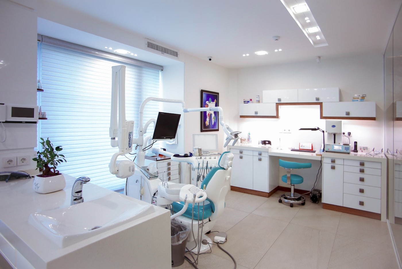 A spotless modern dental operatory with a teal dental chair, overhead examination light, and bright white cabinetry — the chair that needs to stay full