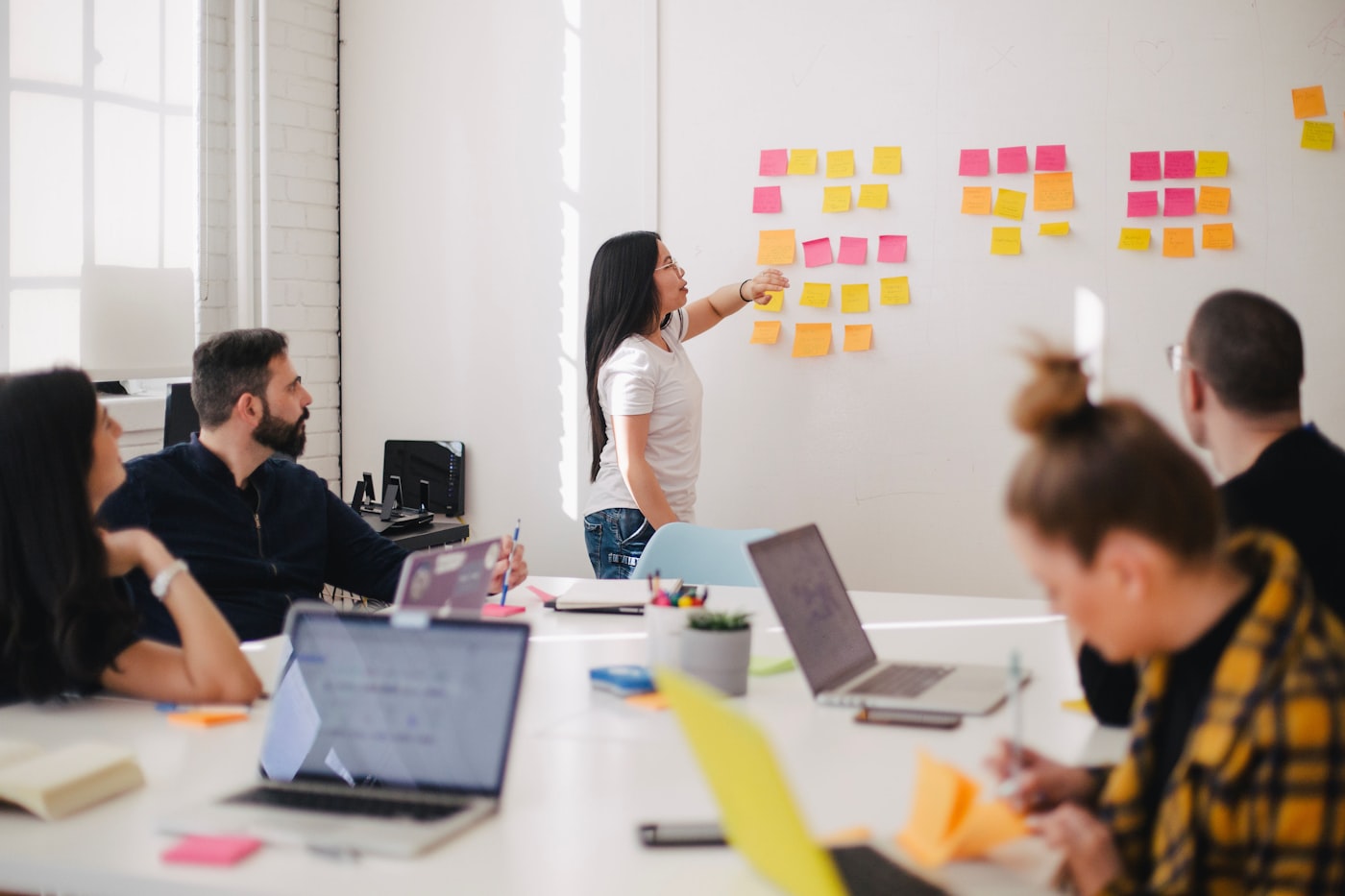 A small growth-stage team collaborating around laptops in a bright modern office, with colourful sticky notes stuck to a whiteboard — tribal knowledge being pulled out of people's heads